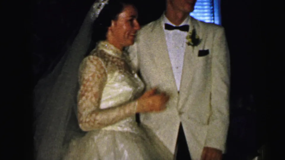 The bride smiles beside the groom at a 1964 Camden, New Jersey wedding reception, with blinds and dark interior visible behind them.