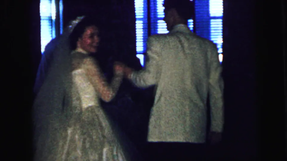 The bride and groom continue their 1964 wedding dance, seen from behind against blue-lit windows in a private reception room.