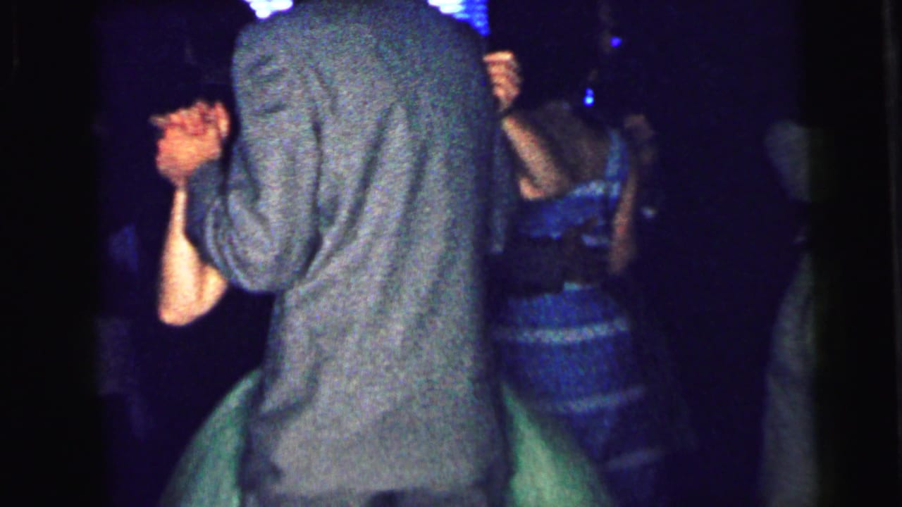 Boy in a gray suit faces away while dancing with a girl in a green dress among other couples.