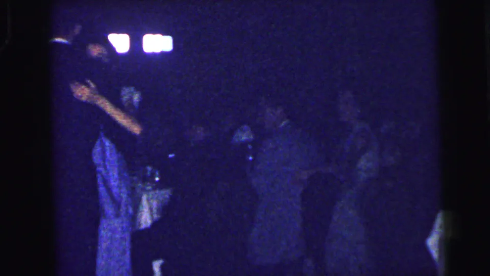 Low-light 8mm frame from the Camden homecoming dance, with figures moving near tables and bright background lights.
