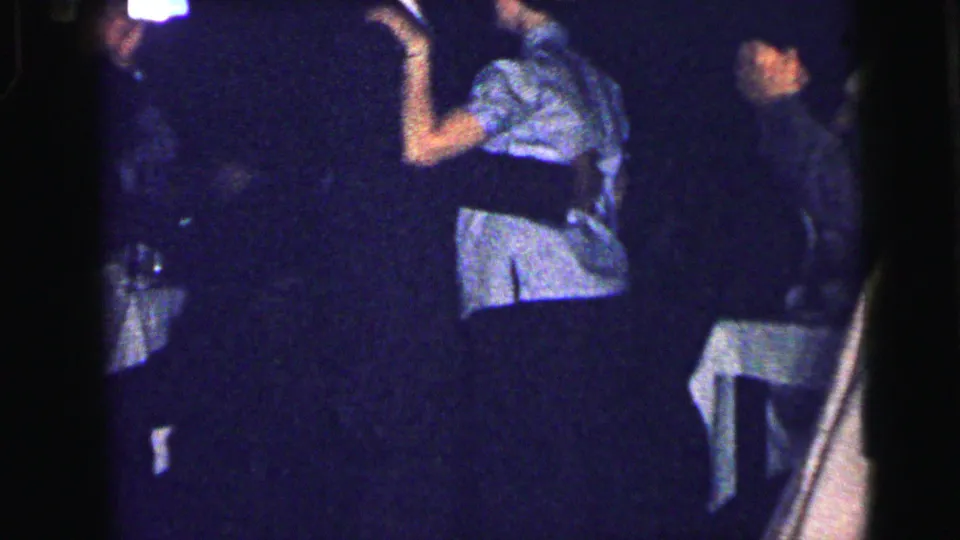 Adults dance closely in a dark Camden interior, including a woman in a light dress and white hat, in 1964 8mm footage.