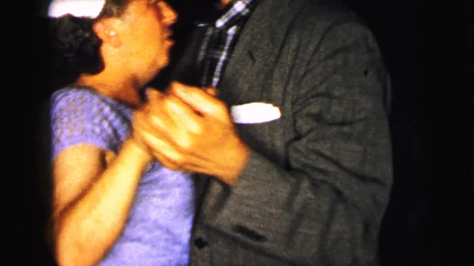 A close 1964 Camden home-movie frame shows a woman in a light dress dancing hand in hand with a suited man.