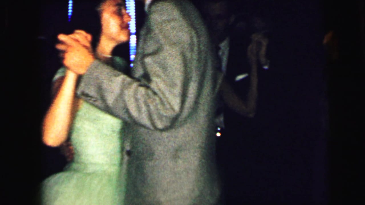 1964 Camden dance scene with a suited man and woman in a pale green dress holding hands beneath dim neon-like lights.