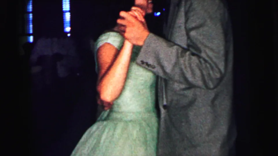 Home-movie frame from 1964 Camden showing a close dance view with a green dress, grey suit, clasped hands, and dark surrounding dancers.