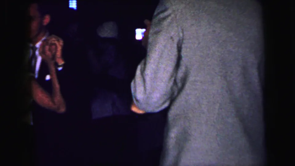 1964 Camden home-movie frame showing adults dancing in a dim venue, with a grey-suited figure from behind and other couples in shadow.