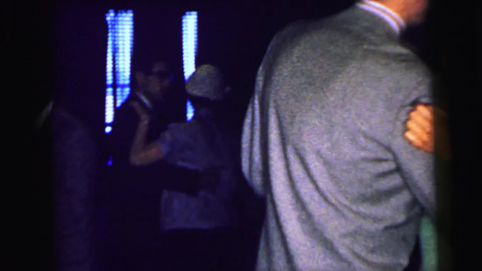 1964 Camden home-movie frame showing a couple dancing in a dim venue with blue vertical lights and other guests nearby.