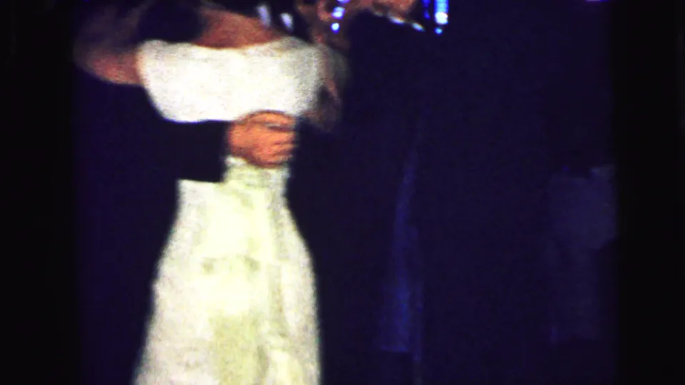 1964 Camden home-movie frame showing a light-colored dress at close range beside dark-suited dancers in a shadowed venue.