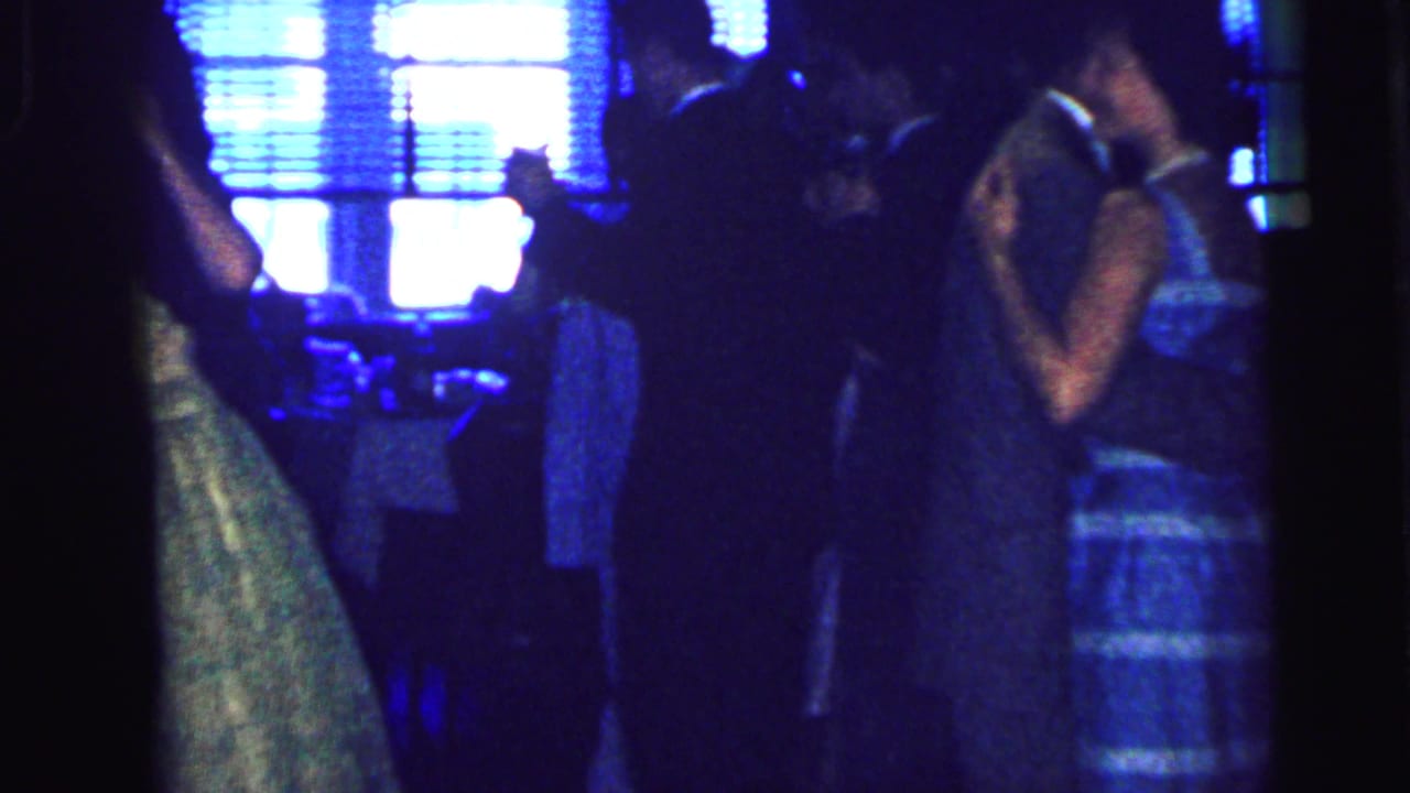 Several couples dance in a dark room with bright window blinds in the background.
