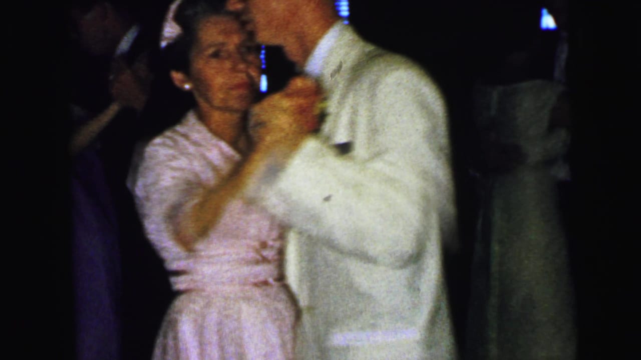 Couple dancing closely at a dim wedding reception, with guests partly visible behind them.
