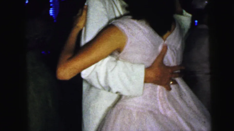 The handheld 8mm frame moves close to a young couple dancing, showing the woman's pale dress and her partner's white jacket.