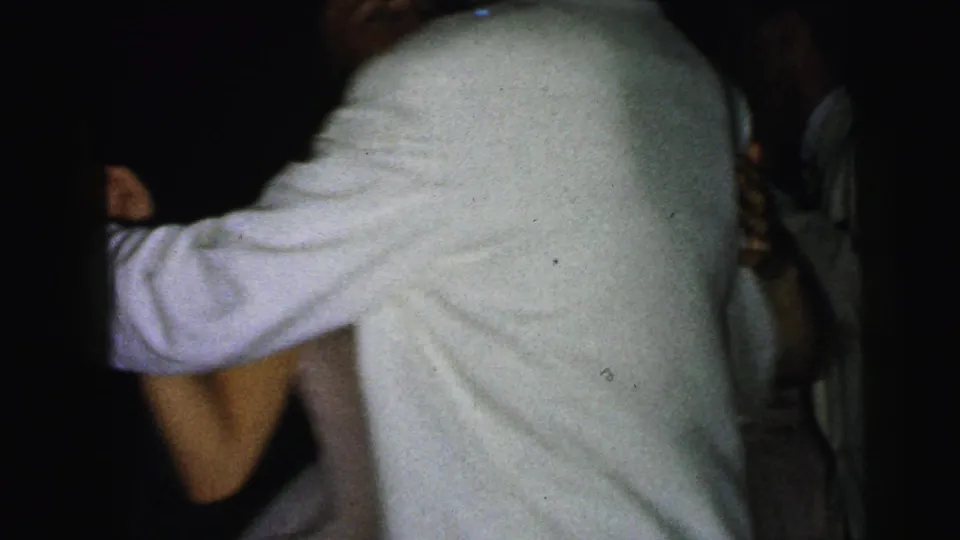 A partner in a white jacket turns or bends during the dance while another figure in formal dress is partly visible at the edge of frame.