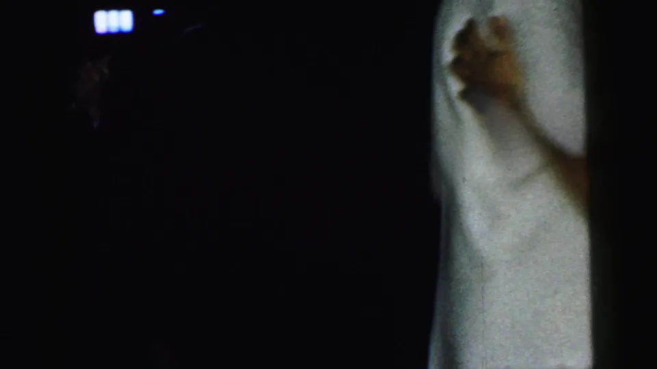 A low-light 8mm frame crops tightly on the dance hold, showing a hand resting on a pale shirt with most of the room in shadow.
