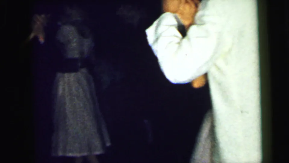 The couple moves toward the right edge of the frame while a shadowed figure in a dress appears deeper in the dim indoor space.