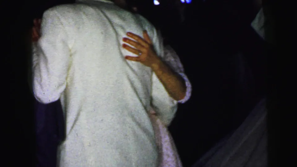 Low-light 8mm frame from the Camden wedding showing the back of a white-suited man dancing with a woman mostly obscured by shadow.