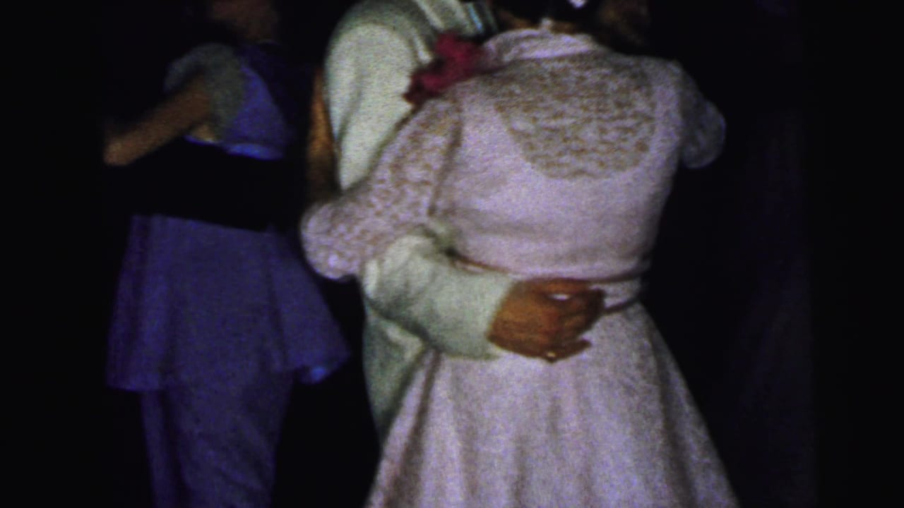 Couple dancing closely in a dim room, with a woman in a light lace dress and other guests nearby.