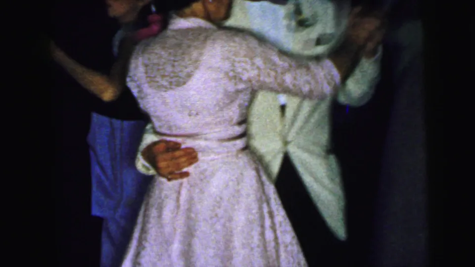 1964 Camden home-movie frame showing a close partner dance, with a light formal dress, lace sleeves, and a man's pale suit jacket.
