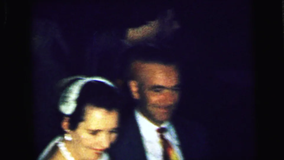 1964 Camden home-movie frame with a suited man and a woman partly visible in low light during the indoor social gathering.
