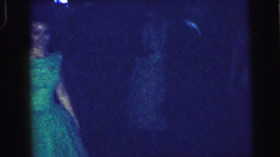 1964 Camden home-movie frame with a woman in a green dress partly seen beneath blue-tinted indoor lighting at the social event.