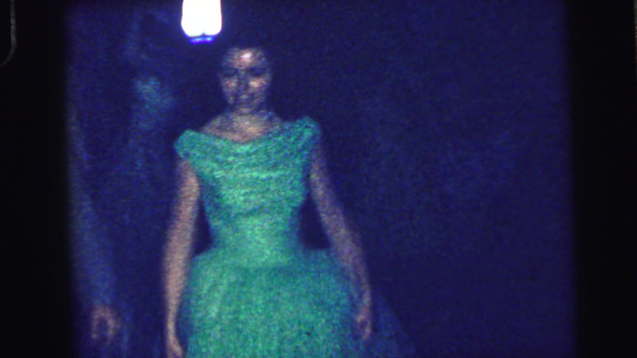 A formally dressed young woman in a green gown walks through a dark Camden, New Jersey venue in 1964 home-movie footage.