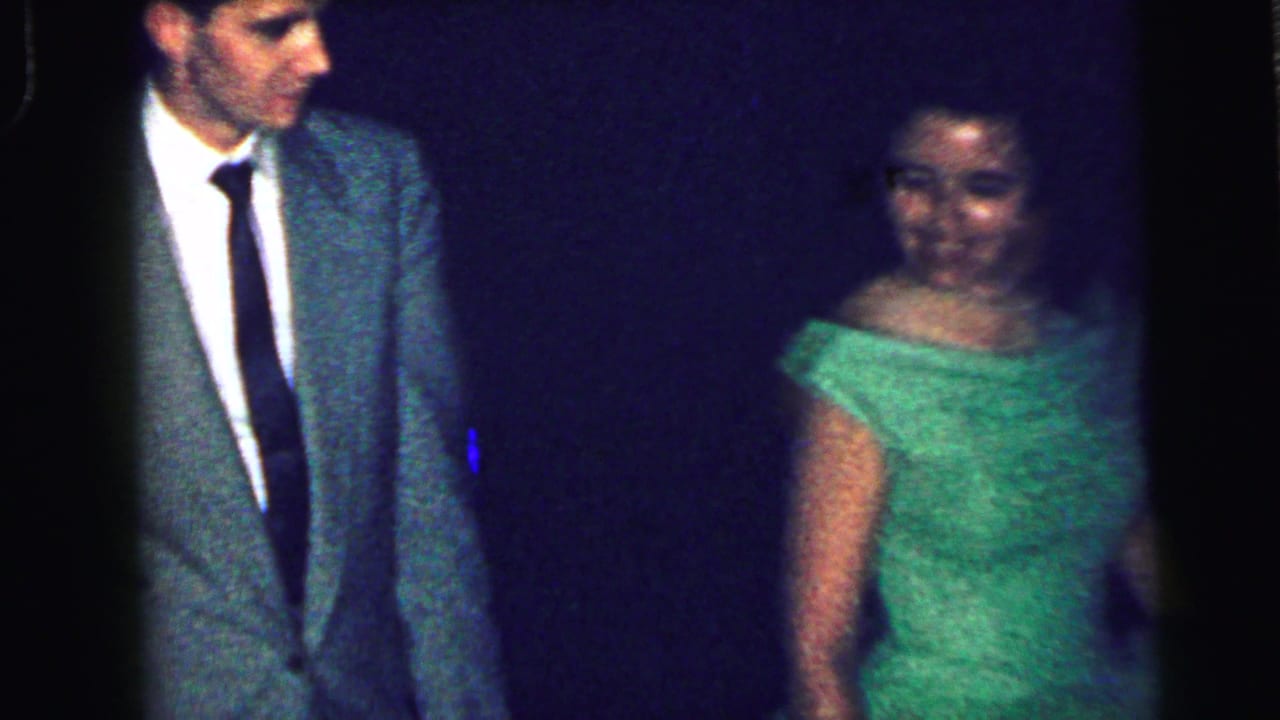A man in a gray suit and tie walks near a young woman in a green gown during the 1964 formal gathering.