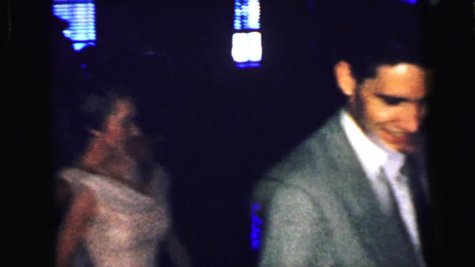 A woman in a pale formal dress appears near a man in a gray suit in grainy 1964 home-movie footage from Camden.
