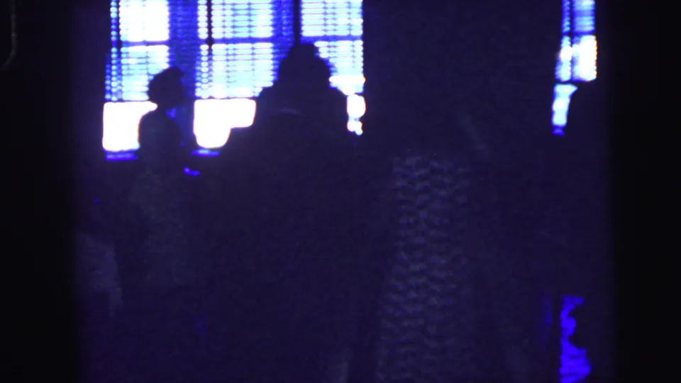 Silhouettes of guests stand against bright blue-lit windows in the formal event venue, with details softened by 8mm grain.