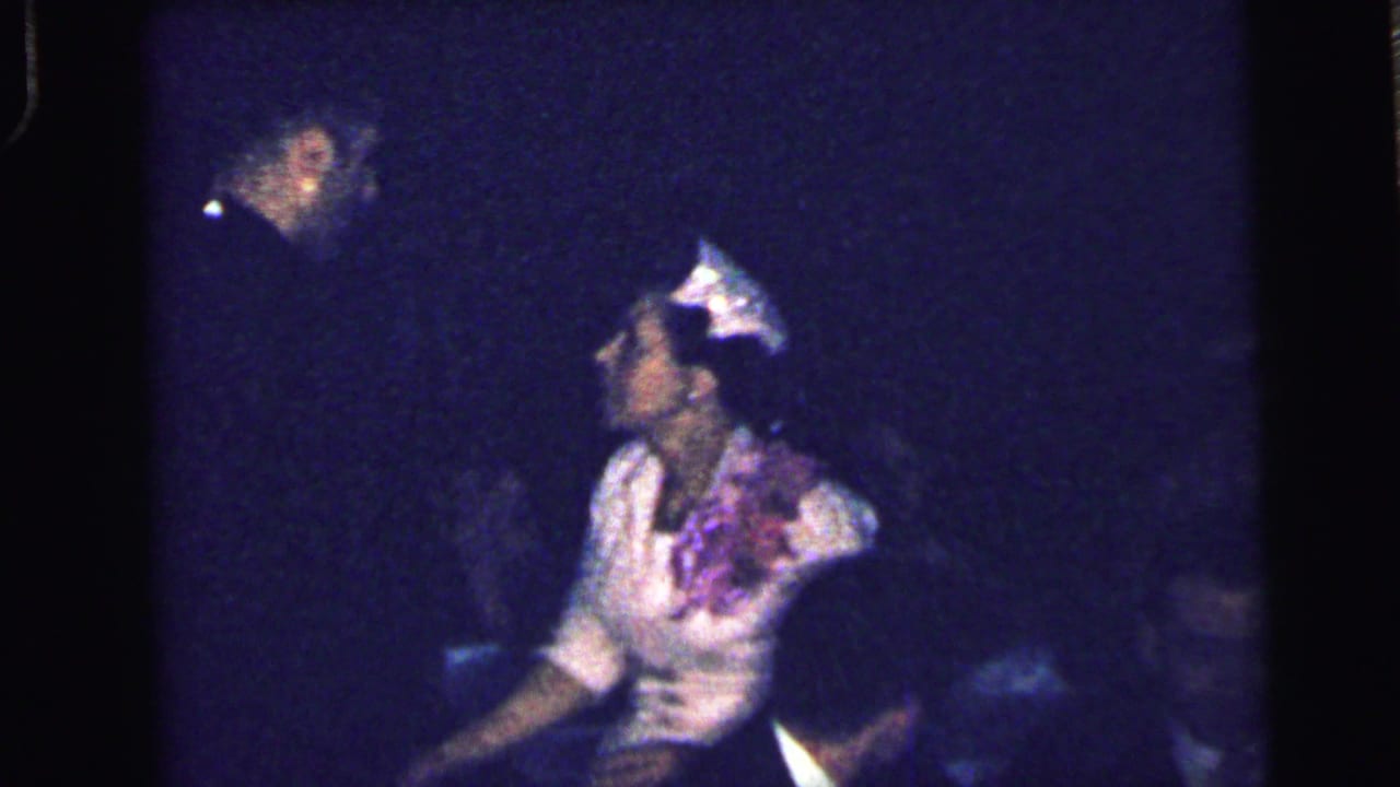 Dim interior with seated adults, including a woman in a light dress and hat near the center.