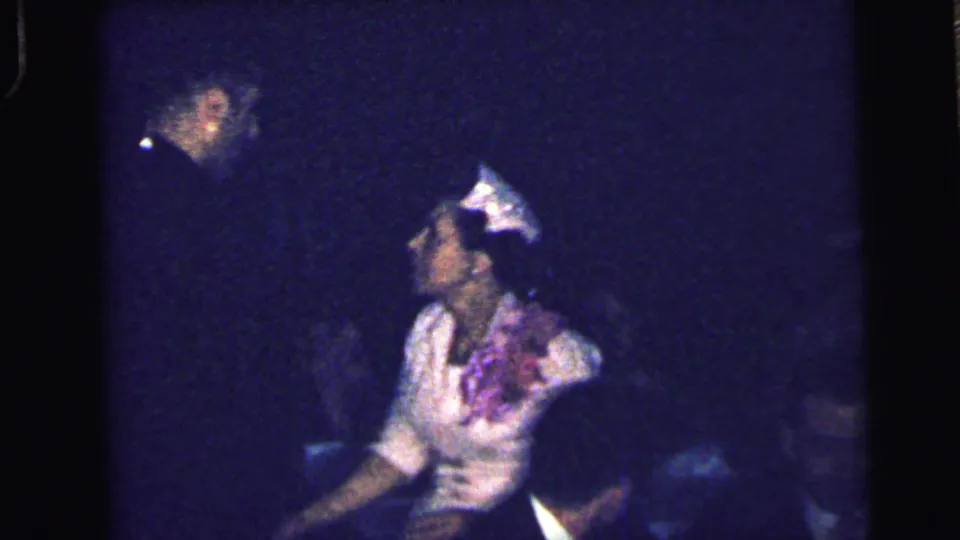 1964 Camden home-movie frame showing adults at a dim formal gathering, with a woman in a light dress and hat seated in profile.