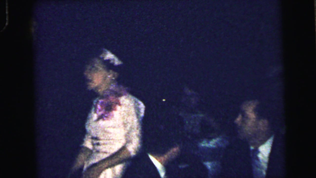Woman in a light dress and hat sits among men in dark suits in a dim room.