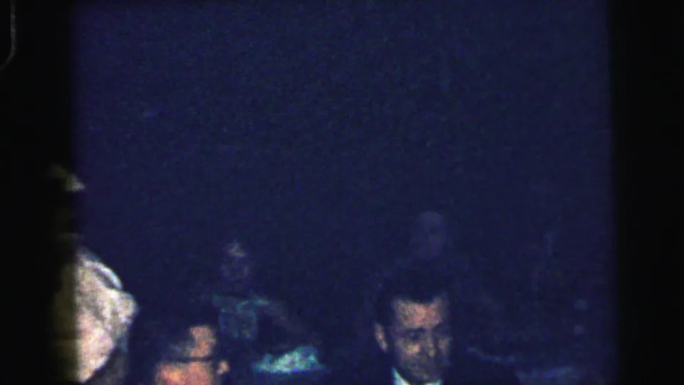 Low-exposure 1964 Camden home-movie frame showing seated adults at a formal indoor gathering, with faces partly obscured by shadow.