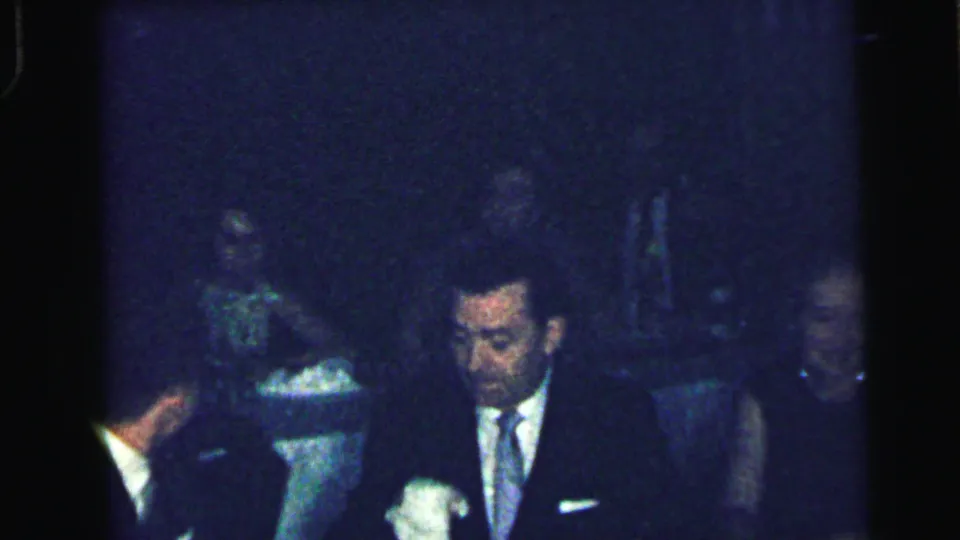 1964 Camden frame showing a suited man seated at a table during a dim formal gathering, with other adults partly visible around him.