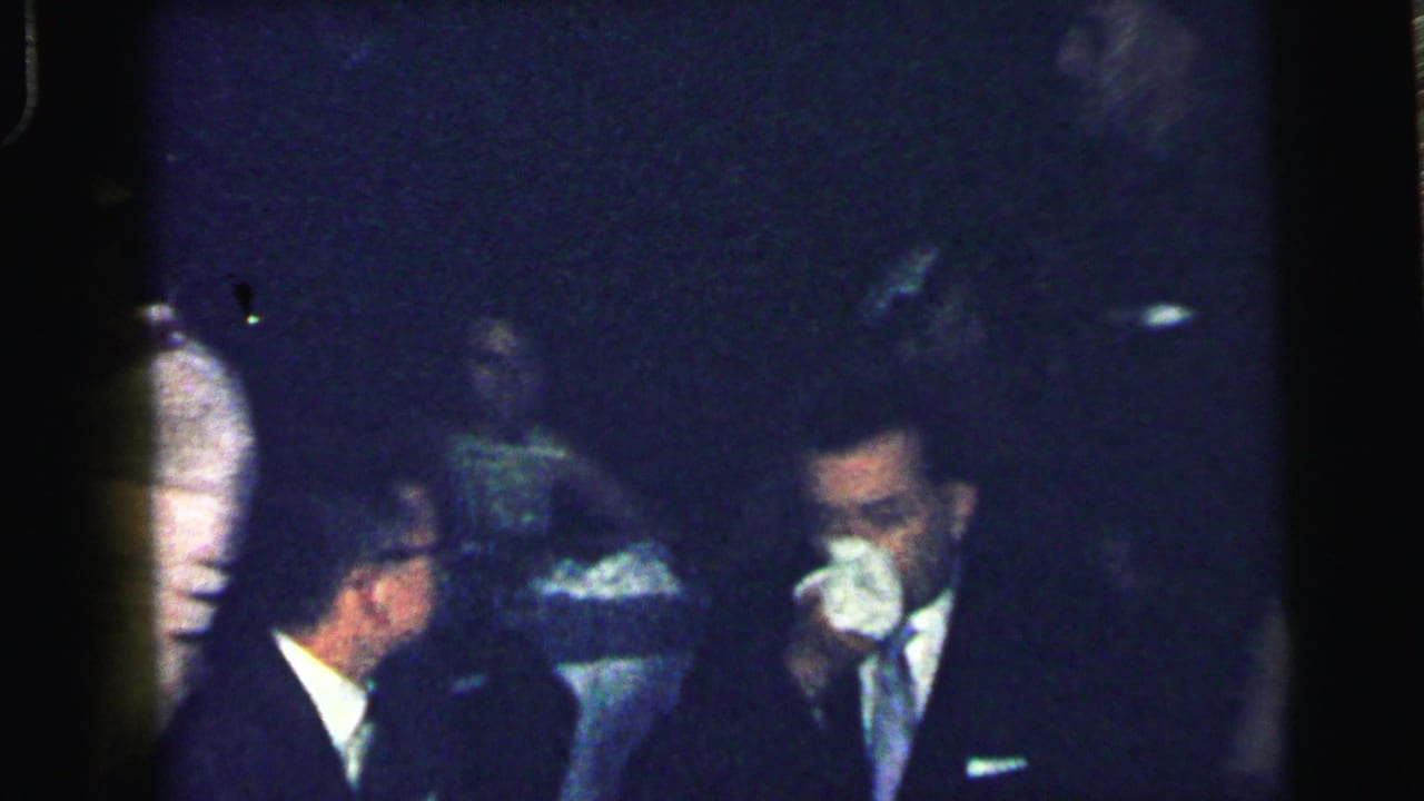 Suited men sit or stand in a dark room while one man holds a white handkerchief to his face.