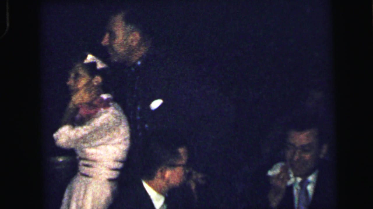 A woman in a pale dress stands near a man while another suited man holds a handkerchief at the right.