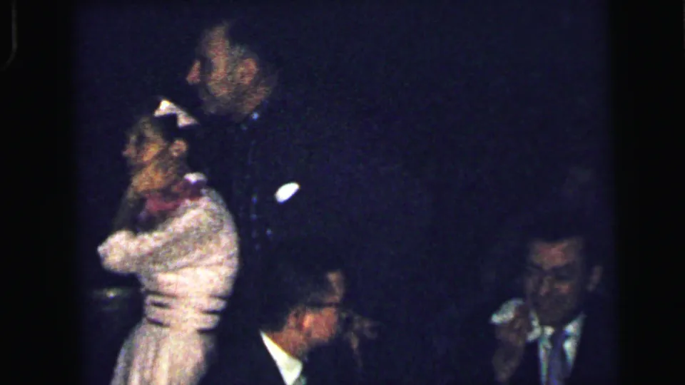 1964 Camden formal-event frame with a woman in a pale dress near a man and a suited guest wiping his nose at the side.