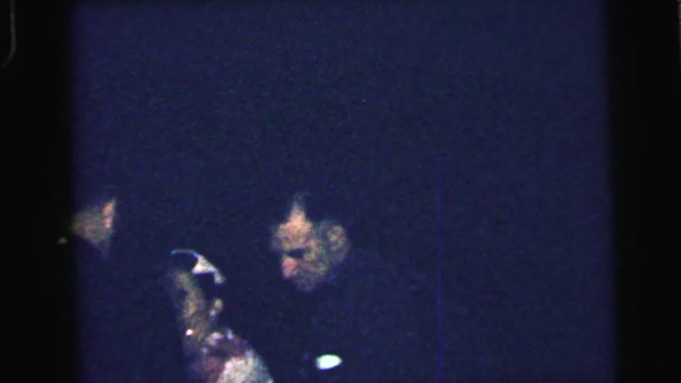 1964 Camden home-movie frame showing a man and woman close together during a dim indoor formal gathering.