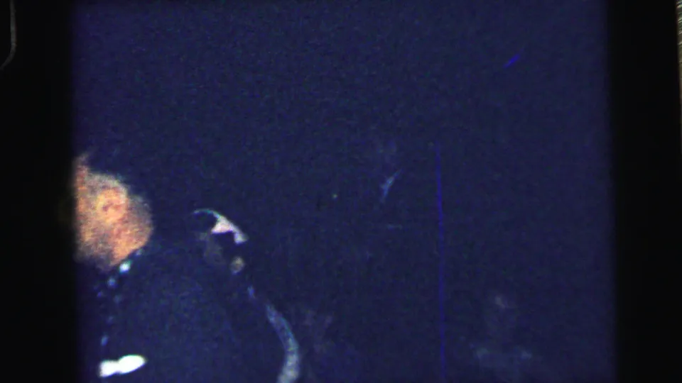 1964 Camden frame with a partly visible suited man at the edge of a dark blue, grainy home-movie scene.