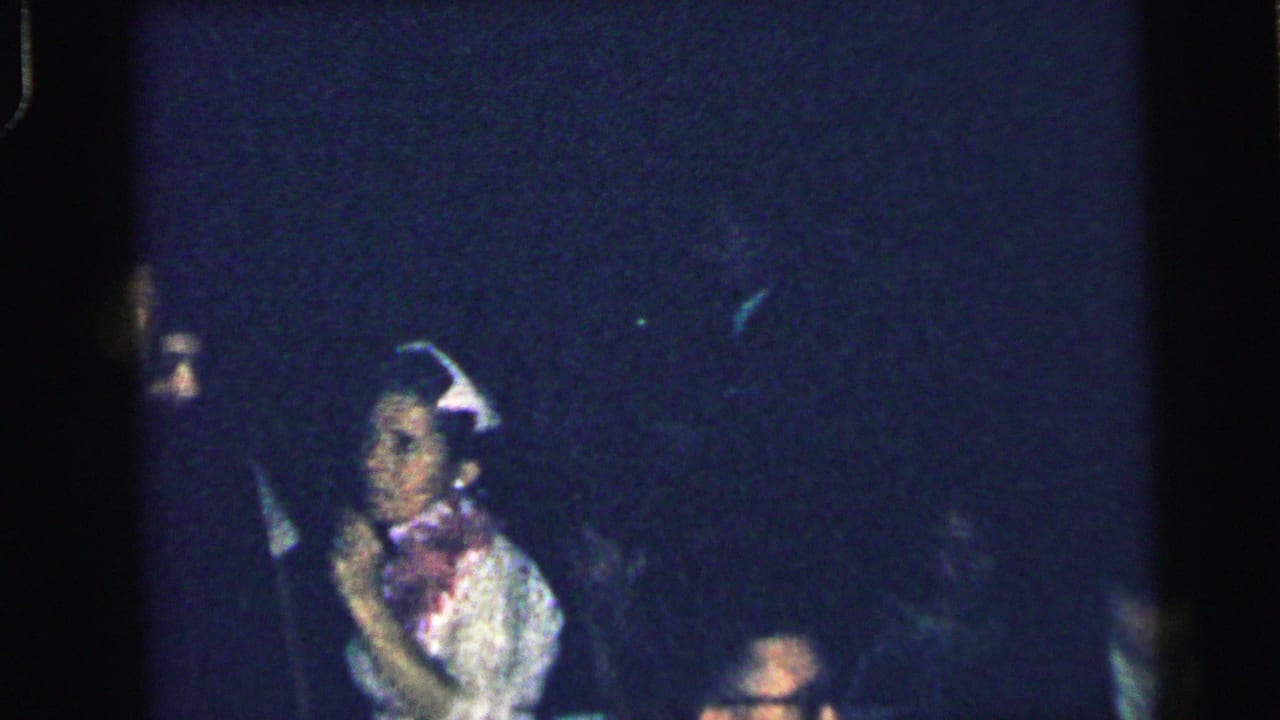 Woman in a light dress and decorative hat stands in a dark party setting.