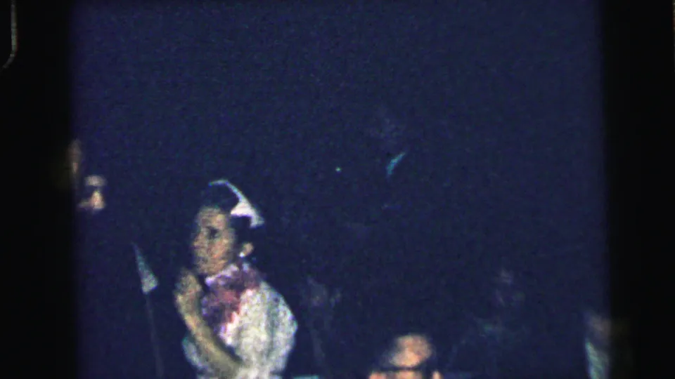 In this 1964 Camden home-movie frame, a woman in a light dress and decorative hat stands amid deep shadows at a social gathering.