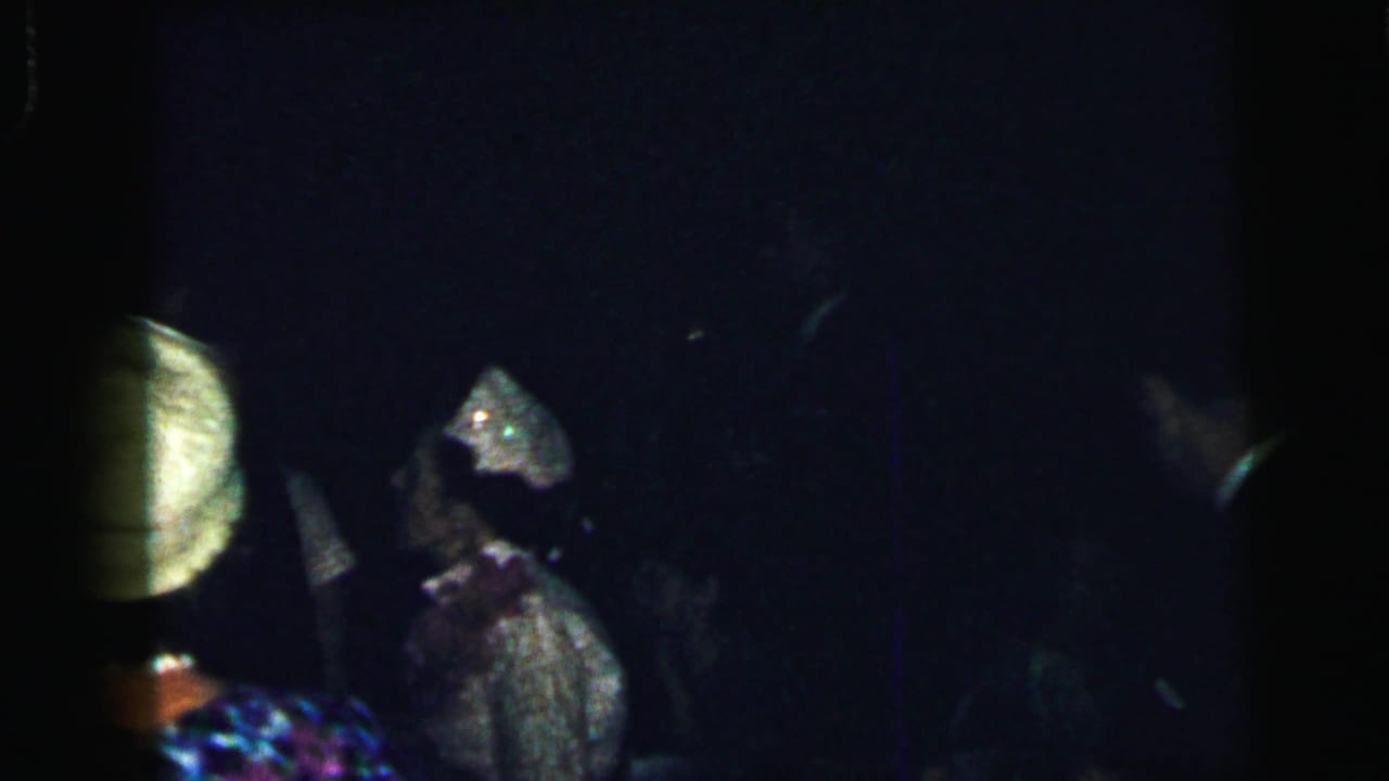 A woman in a light outfit and decorated hat appears in profile while another partygoer stands nearby in the dim Camden gathering.