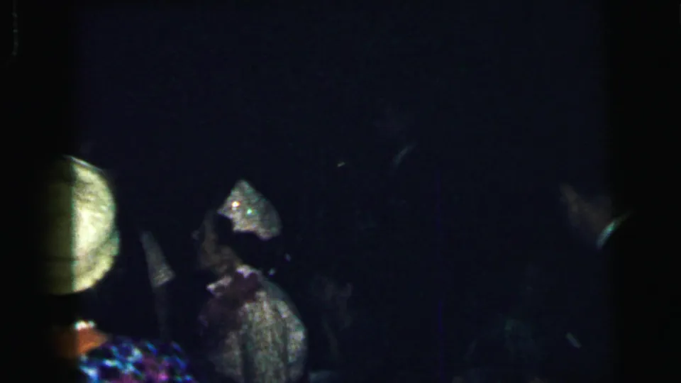 A woman in a light outfit and decorated hat appears in profile while another partygoer stands nearby in the dim Camden gathering.