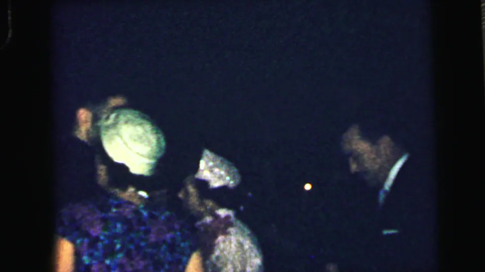 Several adults are partly visible in a low-light 1964 Camden party scene, including a woman with a pale hat and a man in a dark suit.