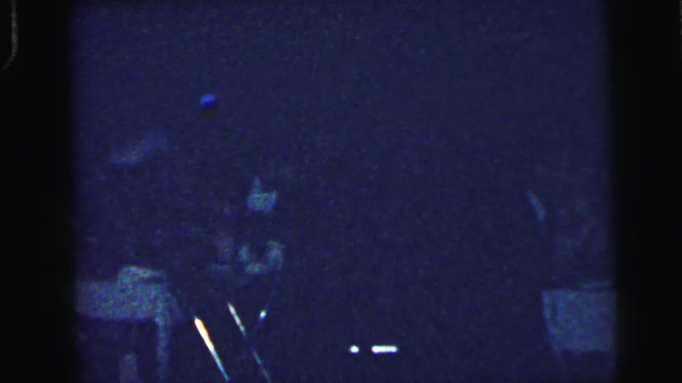 Grainy 1964 8mm frame from a Camden wedding reception showing a dim interior with guests and a small table in view.