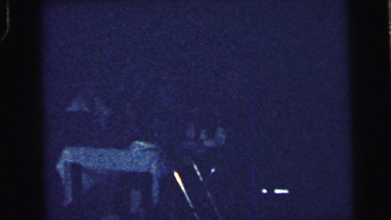 Grainy home-movie frame from the Camden wedding reception with guests around a table in low blue-tinted light.