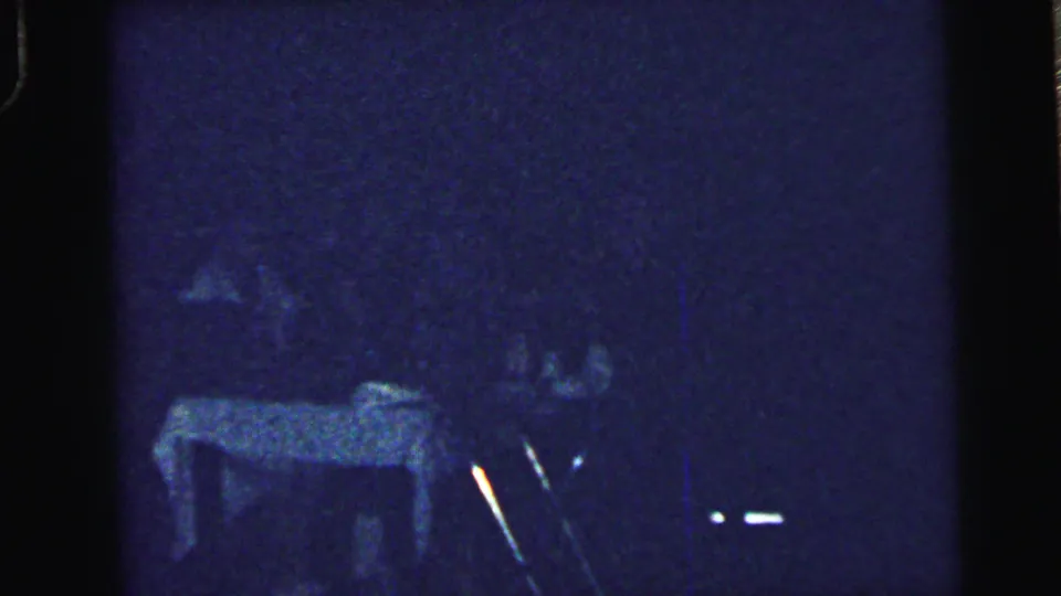 Grainy home-movie frame from the Camden wedding reception with guests around a table in low blue-tinted light.