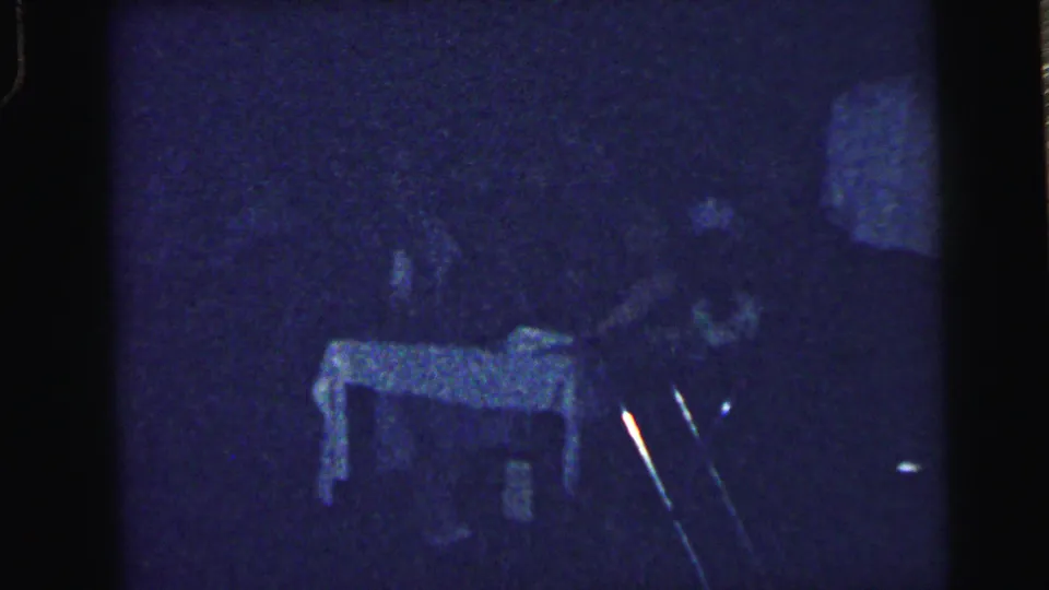 Low-light 1964 Camden wedding reception frame showing a table and indistinct guests through heavy 8mm grain.