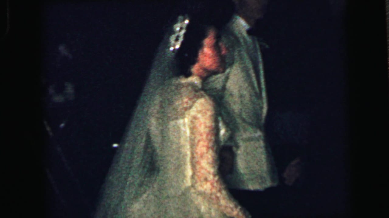 1964 Camden wedding home movie frame showing a bride in a veil leaning toward a groom in a light suit during a dim indoor reception.