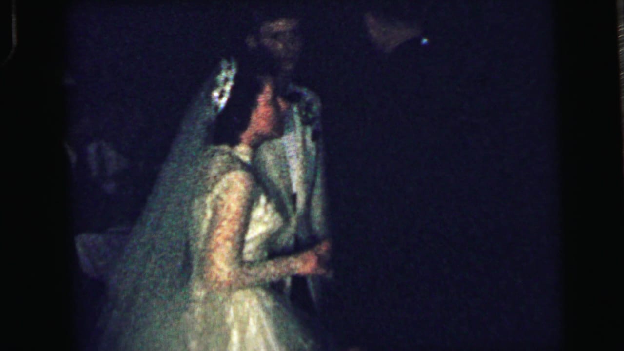 1964 Camden reception frame with the bride in lace sleeves and veil embracing the groom as nearby guests appear in low light.
