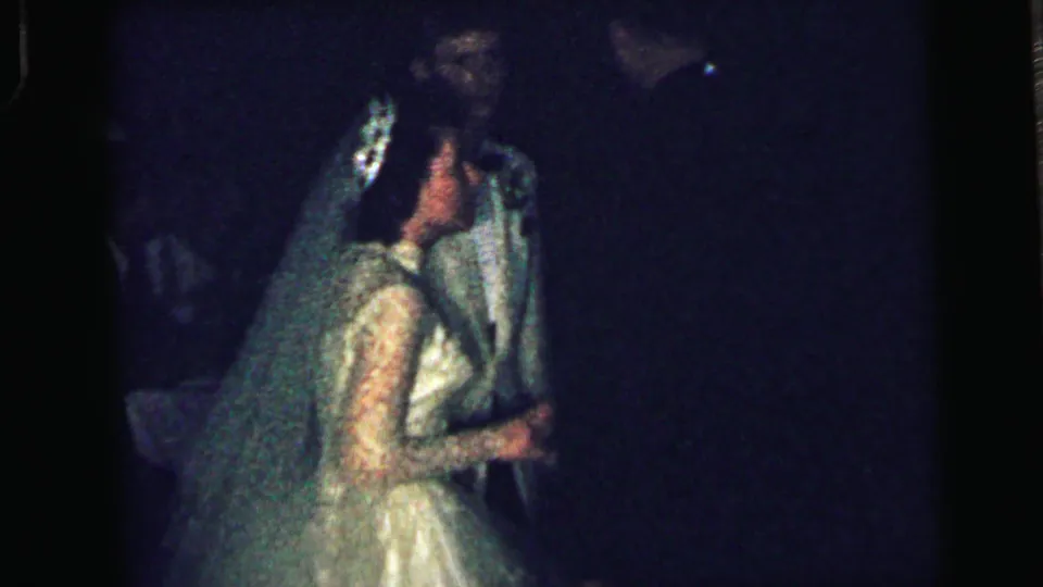 1964 Camden reception frame with the bride in lace sleeves and veil embracing the groom as nearby guests appear in low light.
