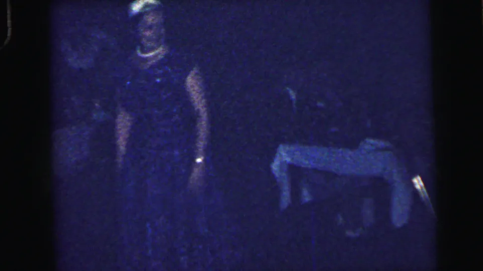 Low-light 1964 Camden reception frame showing a guest in dark clothing near a small cloth-covered table, with the room mostly in shadow.
