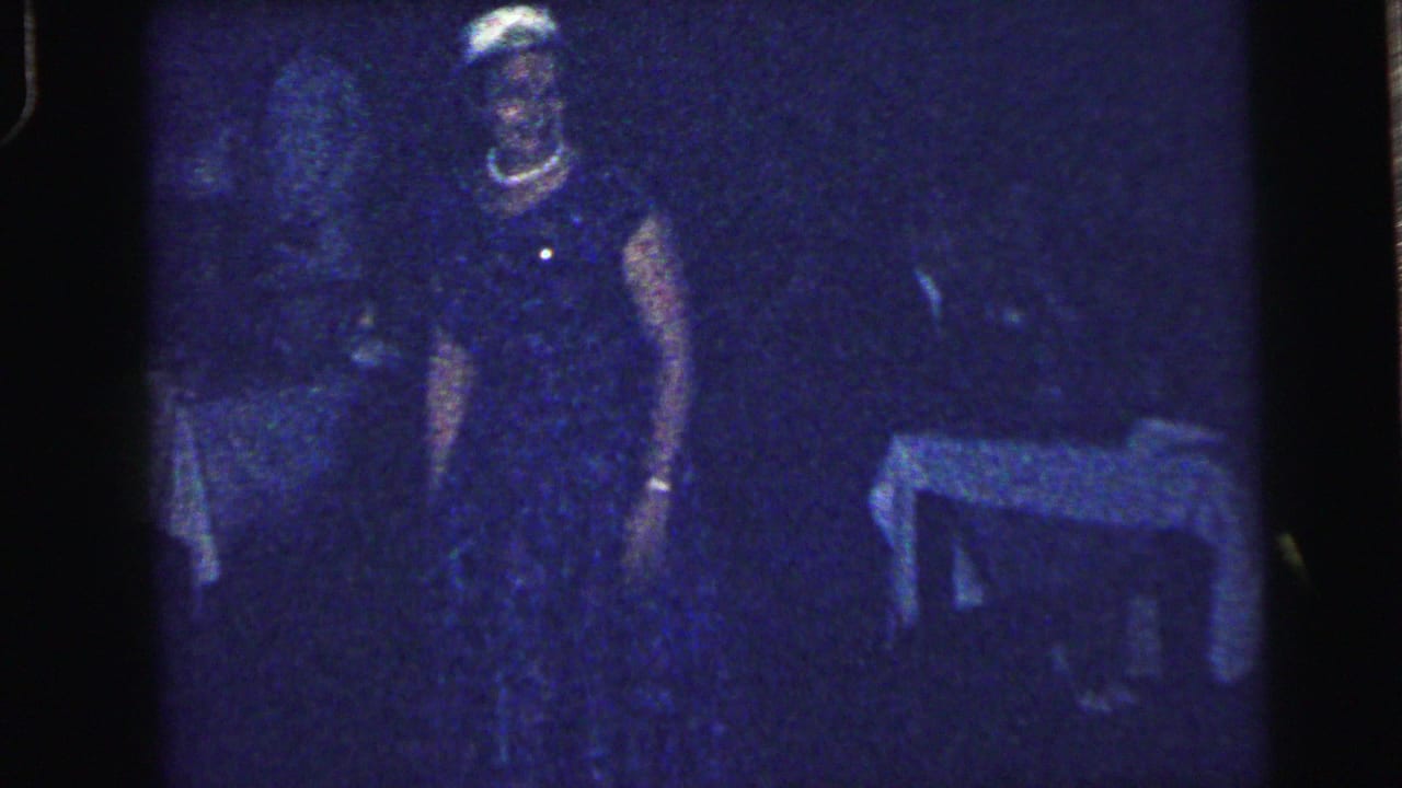 Dim blue-tinted frame with adults standing near a table in a dark room.